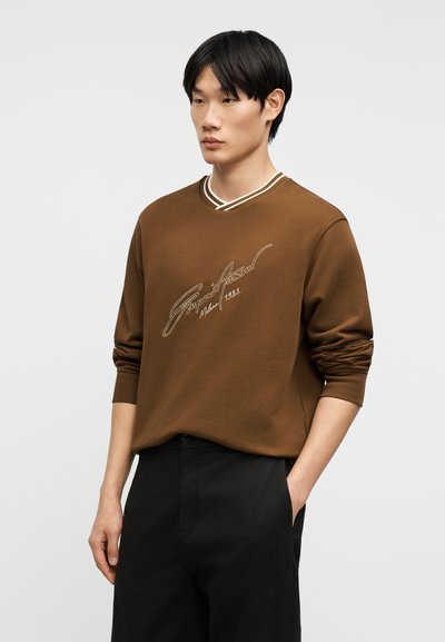 Νεαρός άνδρας με καφέ φούτερ με λευκό κείμενο, μαύρο παντελόνι, στέκεται με το ένα χέρι στην τσέπη μπροστά σε ένα απλό φόντο.