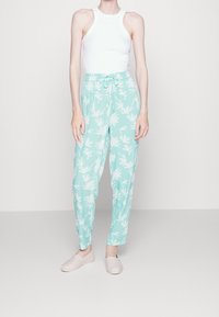 Person som bär en ärmlös vit topp, ljusblå byxor med dragsko och vitt bladmönster, samt blekrosa slip-on skor, står framför en enfärgad bakgrund.