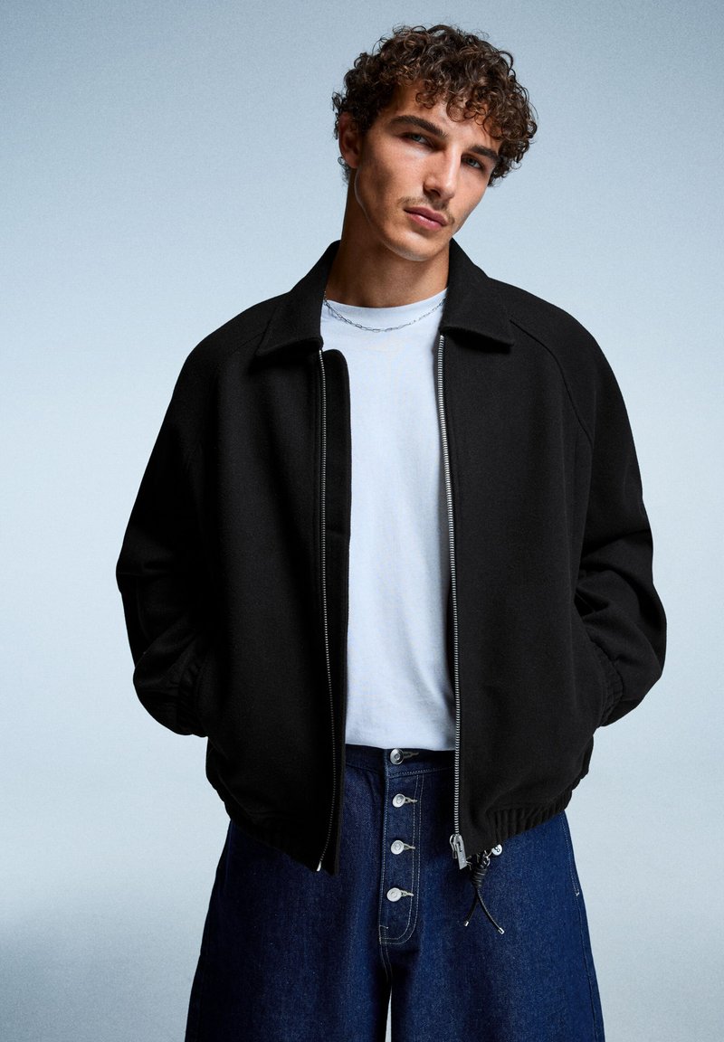 Veste zippée noire avec col montant et poignets côtelés, portée sur un T-shirt blanc, associée à un pantalon en denim bleu taille haute.