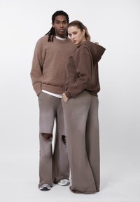 Kaks mudelit kannavad üle mõõdu pruune riideid: karvane kampsun ja kapuutsiga dressipluus, koos laiade jalgadega pükstega, millel on suured rebendid.