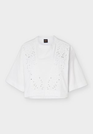 Fehér, rövid ujjú, crop felső széles ujjakkal és virágmintás kivágásos hímzéssel az elülső panelen, egy egyszerű háttér előtt lógva.
