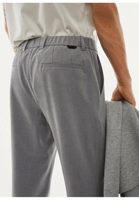 Szare spodnie o gładkiej fakturze, z elastyczną talią i dwiema tylnymi kieszeniami. Szara kurtka przewieszona jest przez ramię noszącego.