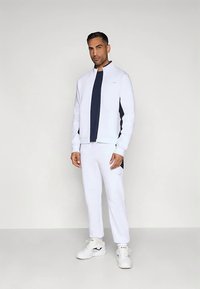 Muškarac stoji u bijelom jaknoma s patentnim zatvaračem i odgovarajućim hlačama, ispod nosi tamnoplavu majicu, a na nogama ima bijele tenisice. Pozadina je jednostavna.