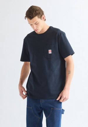 Giovane uomo che indossa una maglietta blu navy con un logo sulla tasca del petto e jeans blu, che guarda verso il basso con le mani vicino ai fianchi.