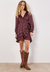 Bordeauxfarbendes Wickelkleid mit langen Ärmeln, gerüschtem Saum und Stern- und Mondmuster. Getragen mit braunen Cowboy-Stiefeln. Angenehm glattes und leichtes Material.