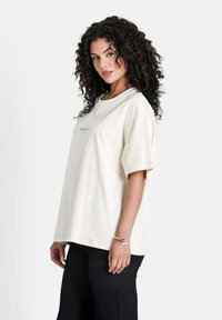 T-shirt oversize beige clair en coton, avec des manches courtes, un col rond et un accent de texte bleu subtil à l'avant. Pantalons texturés noirs.