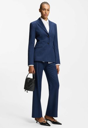 Vrouw gekleed in een marineblauw krijtstreeppak met zwarte kittenhakken, die een kleine zwarte gewatteerde handtas vasthoudt tegen een witte achtergrond.