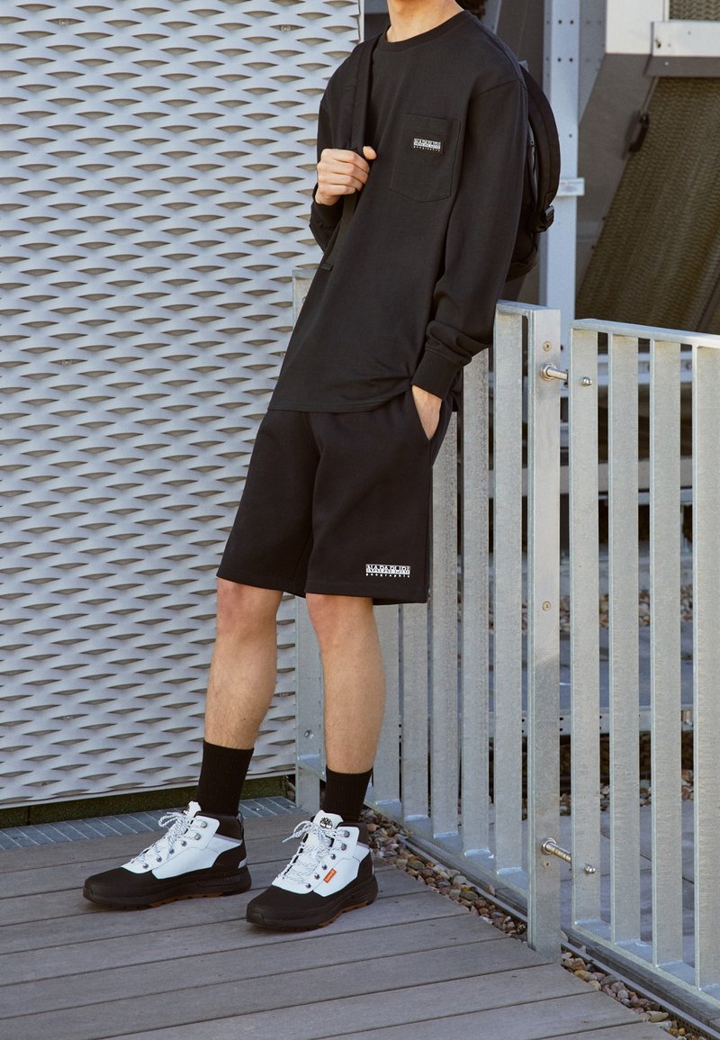 T-shirt noir à manches longues avec poche poitrine, shorts noirs et chaussures de randonnée noir et blanc. Présence d'une marque sur les vêtements et les chaussures.