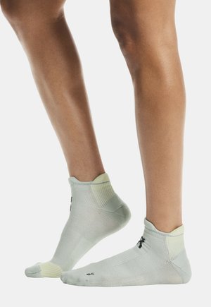 Šviesiai pilkos sportinės kojinės su žemu kulkšnies dizainu, faktūruota medžiaga, geltonos detalės ir juodas logotipas netoli kulno.