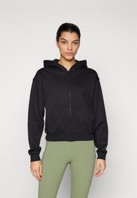 Black zip-up hoodie with a fitted design, large front pockets, and a hood. The fabric has a soft texture, complemented by ribbed cuffs.