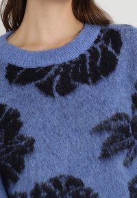 Světle modrý chlupatý svetr s černým vzorem listů, kulatým výstřihem a žebrovaným límcem. Měkká textura dodává vizuální zajímavost.