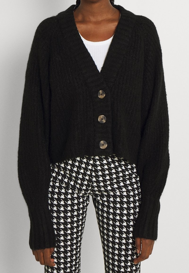 Person wearing a black buttoned cardigan over a white top and black-and-white houndstooth patterned pants, standing against a plain background.