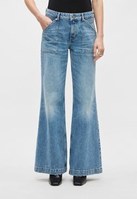 Calças de ganga azul claro com corte evasé, feitas de denim, com cintura alta, grandes bolsos de remendo e textura desbotada. A bainha é crua e desfiada.