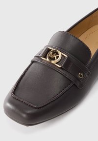 Dark brown leather loafer with a square toe, featuring a gold circular logo and decorative strap across the upper. Smooth texture and stitching detail.