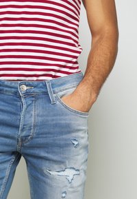 Jeans in denim azzurro chiaro con zone sbiadite e dettagli strappati. Comprendono un passante per cintura e una tasca frontale, abbinati a una maglietta a righe rossa e bianca.