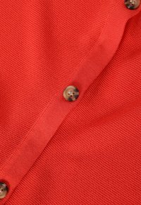 Röd texturerad tyg med en närbild av en knappdetalj som visar en slät, rund form och subtil sömnad.