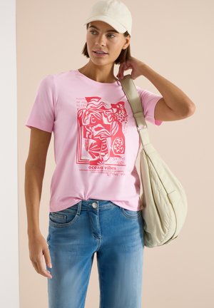 Femme portant un t-shirt rose à motif océanique, un jean bleu, une casquette beige et un grand sac à épaule beige.