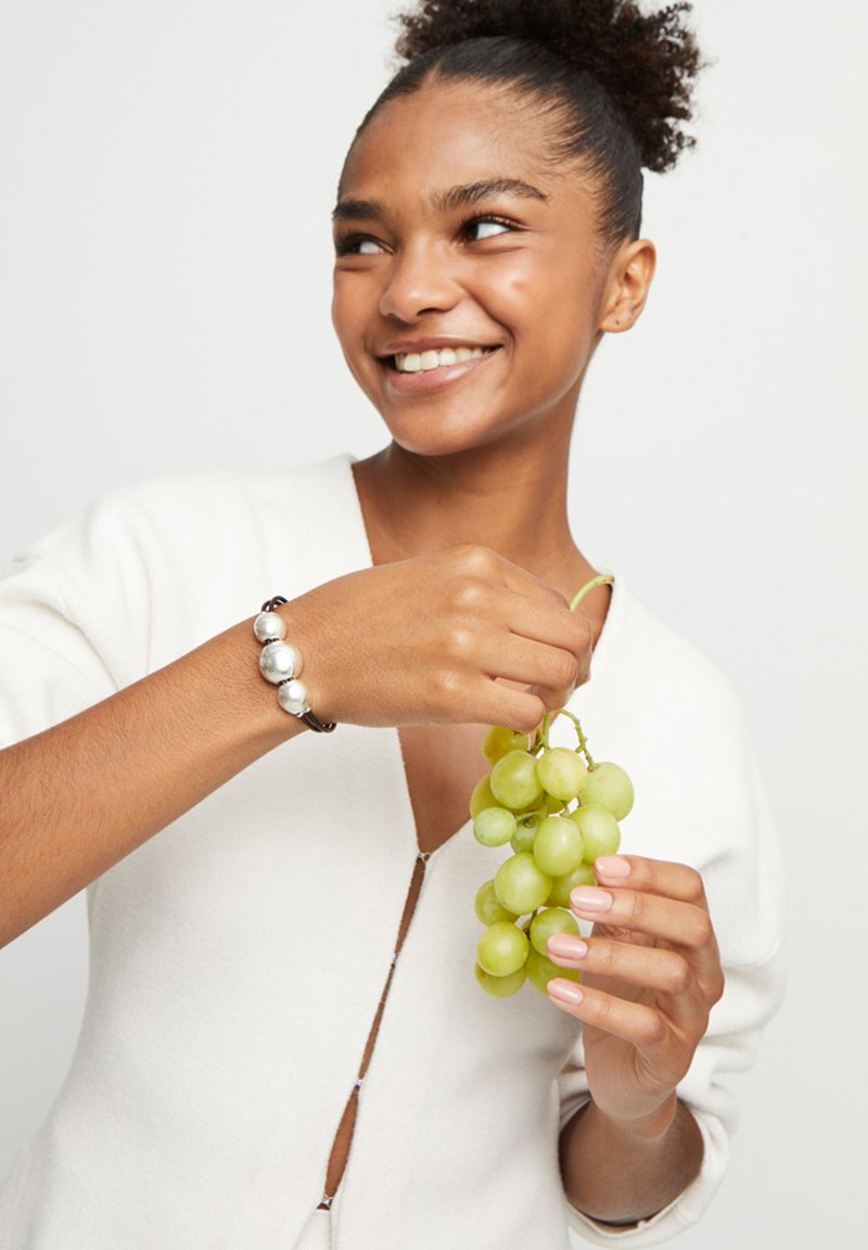 Pulsera de cuentas plateadas en una mano que sostiene un manojo de uvas verdes. La modelo lleva un suéter de color crema con cremallera. Fondo neutro.