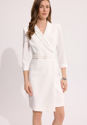 Kobieta ubrana w białą sukienkę z paskiem, rękawami trzy czwarte, kołnierzem z ćwiekami oraz srebrną bransoletką na prawej ręce na jasnym tle.