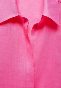 Chemise en lin rose avec col, présentant une texture douce et des coutures visibles. Tombe délicatement, mettant en valeur sa conception légère.