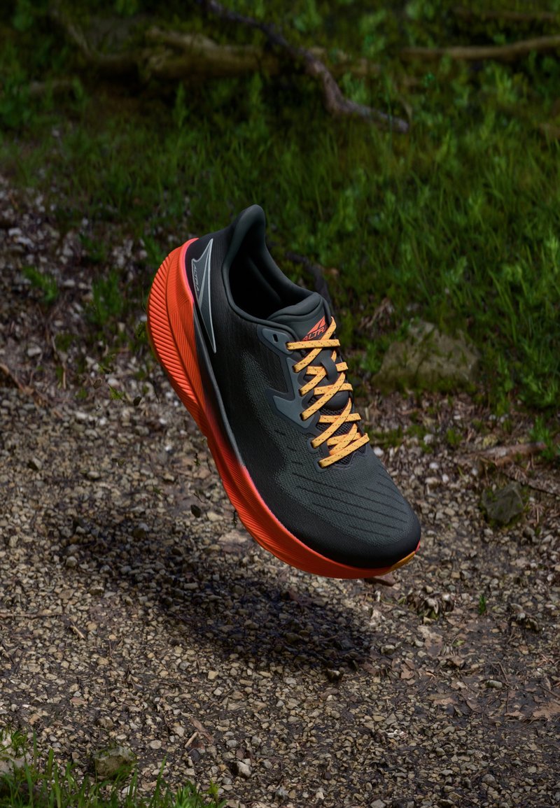Baskets de course noires avec un dessus texturé, lacets orange et semelle extérieure orange vif, présentant un design épuré et une structure de soutien.