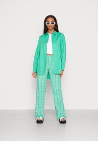 Camisa verde de botão, top cropped branco, calças xadrez verdes e sandálias de plataforma pretas acentuadas com óculos de sol oversized brancos.