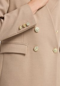 Beiger taillierter Blazer mit glatter Textur, doppelt knöpfbarem Design, goldfarbenen Knöpfen und einer einzelnen Brusttasche.
