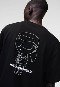 Černé oversized tričko s bílou karikaturní grafikou postavy v slunečních brýlích, pod níž je vytištěno jméno "KARL LAGERFELD".