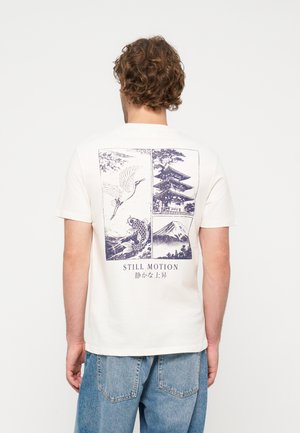 Homme aux cheveux bouclés portant un t-shirt blanc avec une illustration japonaise bleue d'une grue, d'un poisson koi, d'une pagode et d'une montagne, et le texte "STILL MOTION."