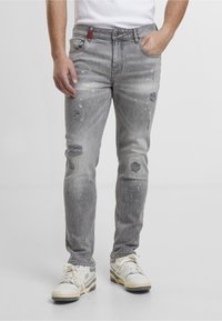 Homme portant un jean gris clair usé avec des éclaboussures de peinture, des baskets blanches et une chemise blanche, debout avec une main dans la poche.