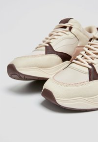 Sneaker beige con accenti marroni. Presenta materiale testurizzato, linguetta imbottita e design con lacci. Suola spessa con pattern di trazione.