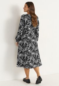 Schwarzes Blumenkleid mit weißen abstrakten Blumenmustern, langen Ärmeln, lockerer Passform und mittellangem Saum. Getragen mit schwarzen Slippers.