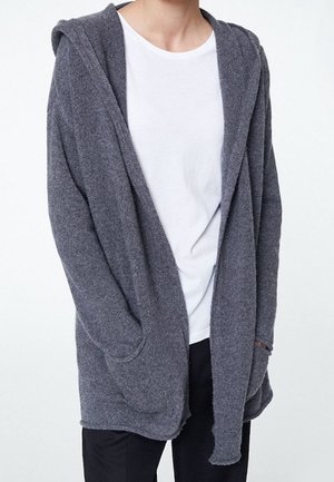 Osoba ubrana w szary kardigan z kapturem, otwarty z przodu, na białej koszulce i czarnych spodniach, stojąca z rękami w kieszeniach.