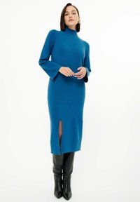 Türkis gestricktes Kleid mit hohem Kragen, weiten langen Ärmeln und einem Oberschenkel-spalt; kombiniert mit knielangen schwarzen, figurbetonten Stiefeln.