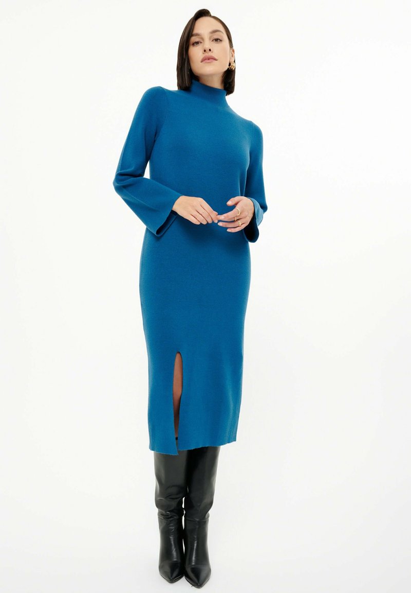 Türkis gestricktes Kleid mit hohem Kragen, weiten langen Ärmeln und einem Oberschenkel-spalt; kombiniert mit knielangen schwarzen, figurbetonten Stiefeln.