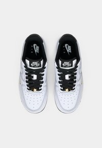 Et par hvite Nike Air Force 1-sneakers med svarte snørebånd, svart innfôring og små gullswoosh-aksenter, sett ovenfra.