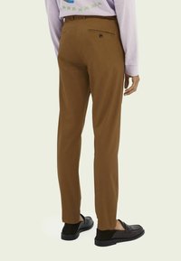 Braune Hose mit geradem Schnitt, mit einer Gesäßtasche und Knopfdetail. Kombiniert mit schwarzen Slippern.