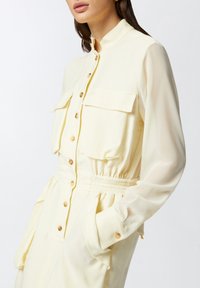Abito button-up giallo chiaro con maniche lunghe, taschini sul petto, vita elastica e dettagli con bottoni. Tessuto morbido e leggero.