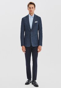 Granatowy żakiet z fakturą z dwiema kieszeniami, jasnoniebieska koszula i ciemne spodnie. Czarne buty eleganckie dopełniają strój. Chusteczka w kieszeni w wzór.