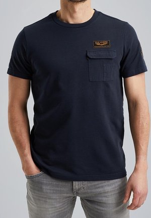 Homme portant un t-shirt bleu marine avec une poche poitrine et une étiquette, associé à un jean gris clair, debout avec une main dans la poche.