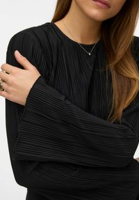 Žena nosi crnu plisiranu bluzu dugih rukava, srebrnu ogrlicu s malim privjeskom i prstenje na lijevoj ruci dok drži desnu ruku.