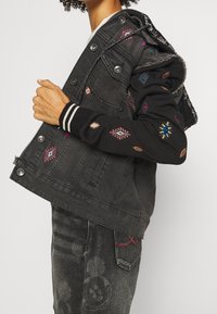 Svart denimjacka med svart huvtröja, prydd med färgglad broderi, dragkedja framtill och elastiska cuffs, som ger en lager-på-lager-look.