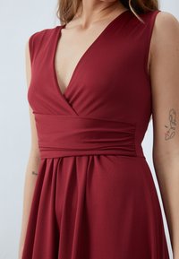 Burgundy mekko syvällä V-päänteellä, tiukasti istuva yläosa ja virtaava pidentävä hame. Valmistettu pehmeästä kankaasta, jossa on kietoutuva vyötärösuunnittelu.