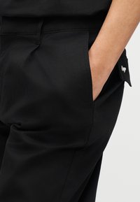 Μαύρο παντελόνι από λείο ύφασμα, με πλαϊνή τσέπη, ραμμένες ραφές και μικρό λογότυπο στην τσέπη. Απλός σχεδιασμός και ουδέτερο χρώμα.