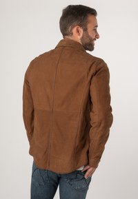 Bruine suède jas met een gestructureerde pasvorm, accentstiksels en een kraag, gecombineerd met blauwe denim jeans. Soepele textuur, minimalistisch ontwerp.