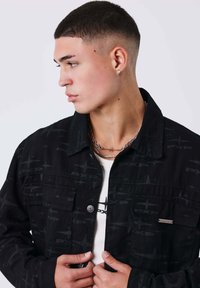 Chaqueta de mezclilla negra con un patrón texturizado, cierre de botones y dos bolsillos delanteros. Usada sobre una camiseta blanca con logo impreso.