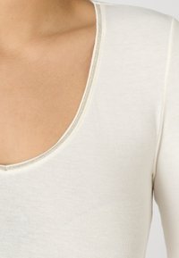 Langarmtop in crème, met een diepe V-hals en een strak aansluitende ribbeltextuur. Afgewerkt met een nette stiknaad bij de hals.