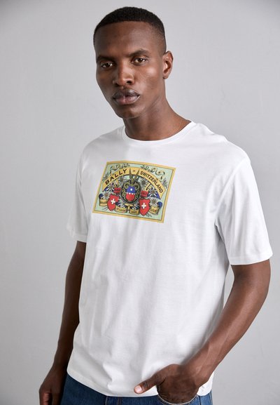 Camiseta de algodón blanca con un estampado gráfico colorido que presenta escudos de armas y el texto "BALLY de SUIZA" en el centro. Corte casual.