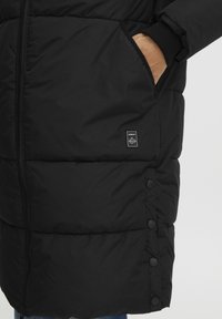 Sort pufferjakke med quiltet design, der har lynlås lukning, lommer i siderne og et lille mærke nær kanten.