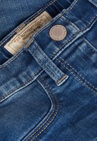 Džínsy z denimu s tmavomodrou prateľnou úpravou, viditeľným šitím, kovovým zapínaním na gombík a štítkom s nápisom "THE AUTHENTIC CUT" v béžovom písme.